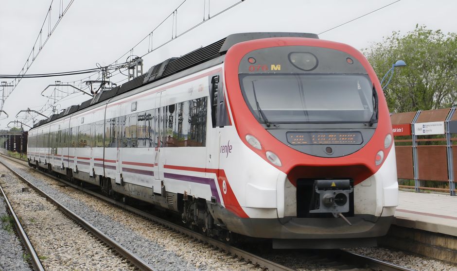 Close-up view of a series of train doors with reflective glass panels, set within a metallic frame, lined up along a train platform. The platform surface features a textured, multi-colored tactile pav